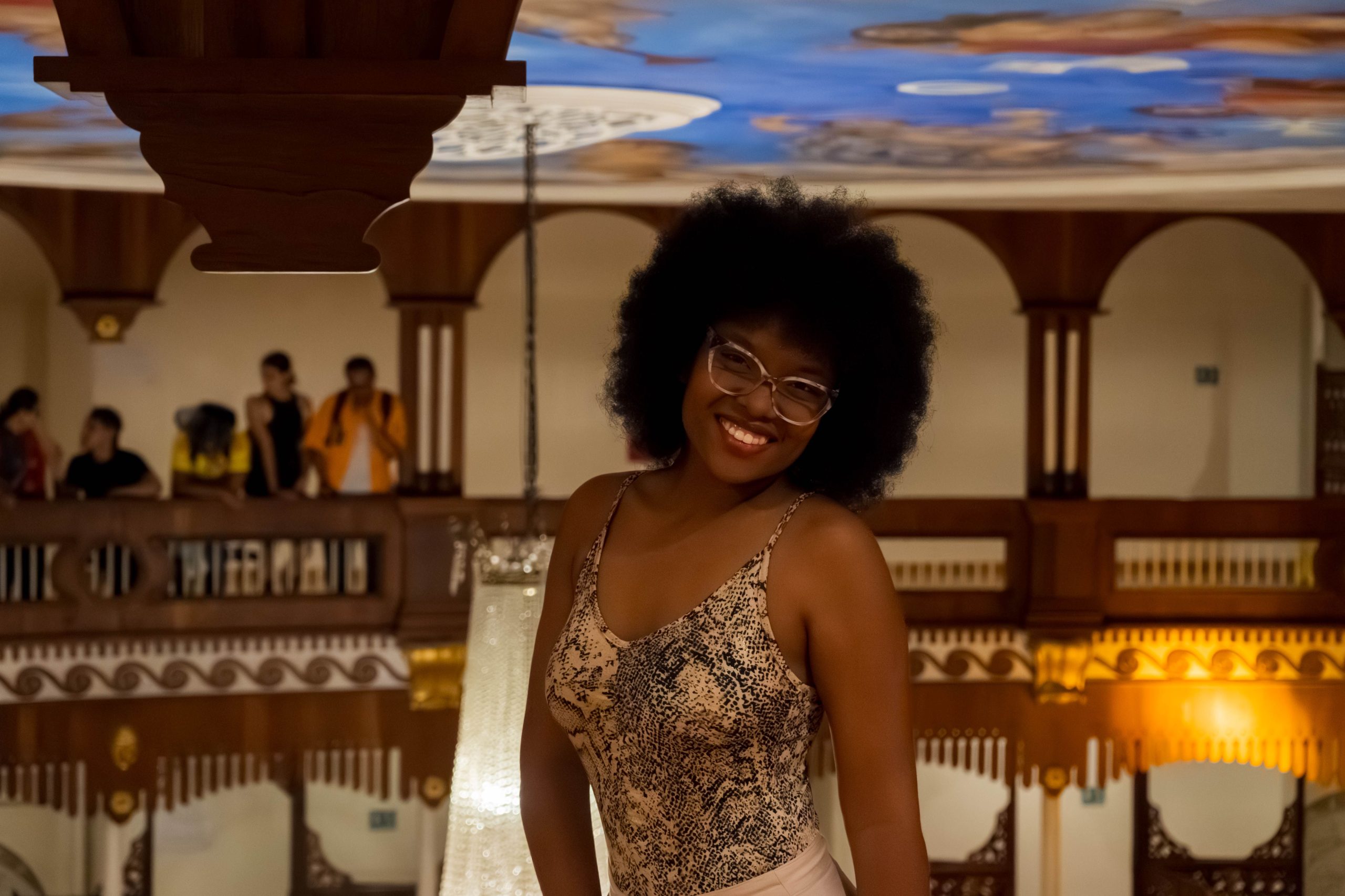 A joyful woman posing indoors with a decorative ceiling and people in the background.